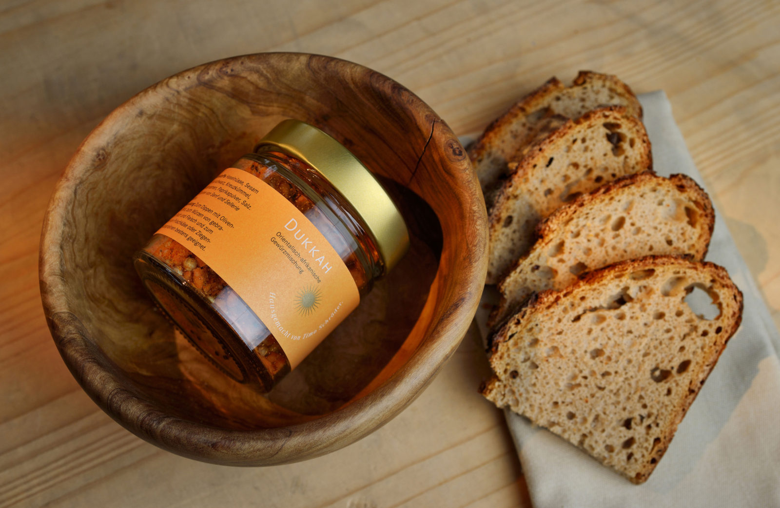 Dukkah Gewürzmischung in einem Glas in einer Holzschale, daneben vier Scheiben Brot auf einer Servierte